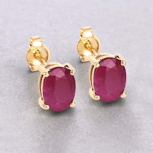 May include: A pair of gold earrings with oval-shaped ruby gemstones. The earrings have a simple stud design.