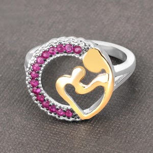 Op de afbeelding: Zilveren ring met een gouden hartvormig ontwerp met een moeder en kind. De buitenste cirkel van de ring is versierd met kleine, ronde, roze edelstenen. Een mooi sieraad.