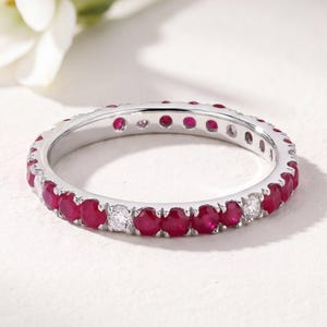 May include: A silver-toned eternity band featuring a repeating pattern of round-cut red rubies and clear white diamonds set in a continuous metal channel. The ring displays a polished metallic finish with alternating gemstone colors.