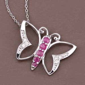 Pode incluir: Um colar com um pingente de borboleta de prata com uma fileira de pedras preciosas rosa no centro e pedras preciosas transparentes ao redor das bordas.