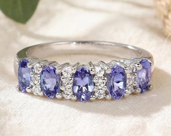 May include: A silver ring featuring a row of oval-cut, light purple gemstones, interspersed with smaller, round, clear stones. The ring's band is a polished silver color, and the gemstones are set close together. The ring is displayed on a white surface.