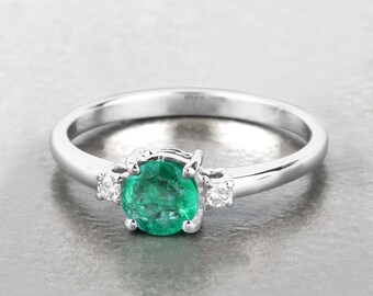 May include: A silver ring with a large, round, bright green emerald as the central stone, with two smaller, clear accent stones. The ring has a polished band, and the gemstones are set in a classic design. A piece of jewellery.