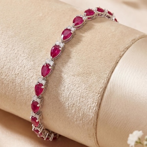 May include: A silver bracelet featuring pear-shaped, deep red gemstones. Each gemstone is set in a silver frame and is interspersed with small, clear accent stones. The bracelet is displayed on a beige surface.