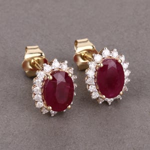 May include: A pair of gold stud earrings featuring oval-cut, deep red gemstones surrounded by a halo of small, sparkling white stones. The earrings are set against a neutral gray background.