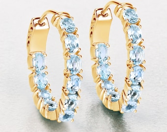 May include: A pair of gold hoop earrings with a row of light blue oval-shaped gemstones around the hoop.