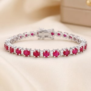 May include: A silver bracelet featuring a series of oval-cut, vibrant red gemstones, interspersed with smaller, clear stones. The bracelet is secured with a clasp and rests on a cream-colored surface, showcasing its elegant design.