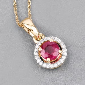 May include: A gold pendant necklace featuring a round, deep red gemstone encircled by a halo of small, sparkling diamonds. The pendant is suspended from a delicate gold chain, creating an elegant and eye-catching piece of jewelry.