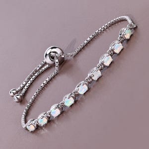 May include: A silver bracelet featuring a row of oval-shaped opals. The bracelet has a delicate chain design and an adjustable sliding clasp. The opals have a milky white color with subtle flashes of iridescence, set in a silver metal.