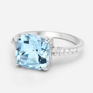 May include: A silver ring with a large, light blue square-shaped gemstone and a row of smaller clear gemstones on the band.