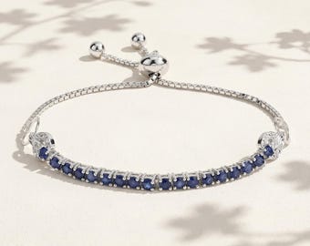 May include: A silver-toned bracelet with a row of small, dark blue gemstones. The bracelet has a sliding clasp for adjustable sizing. The design includes small, clear accent stones at each end of the gemstone row. The background is a soft, neutral color.