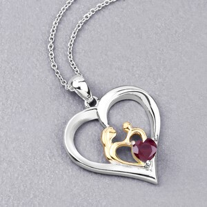 May include: A silver and gold heart-shaped pendant necklace with a red gemstone in the center. The pendant features a design of two figures holding hands inside a smaller heart.