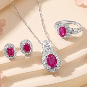 May include: A jewelry set featuring a ruby gemstone. The set includes a pendant necklace, a ring, and a pair of earrings. Each piece has a silver setting and is adorned with a large, oval-shaped ruby surrounded by smaller, clear gemstones.