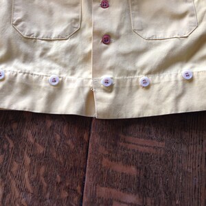 May include: A yellow button-down shirt with white buttons. The shirt has a pocket on the left side and a row of buttons down the front.