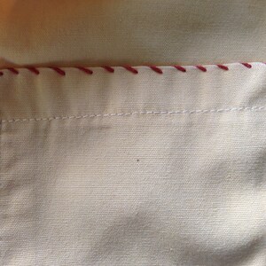 May include: A close-up of a beige fabric pocket with a red stitched border. The pocket is sewn onto a larger piece of beige fabric.