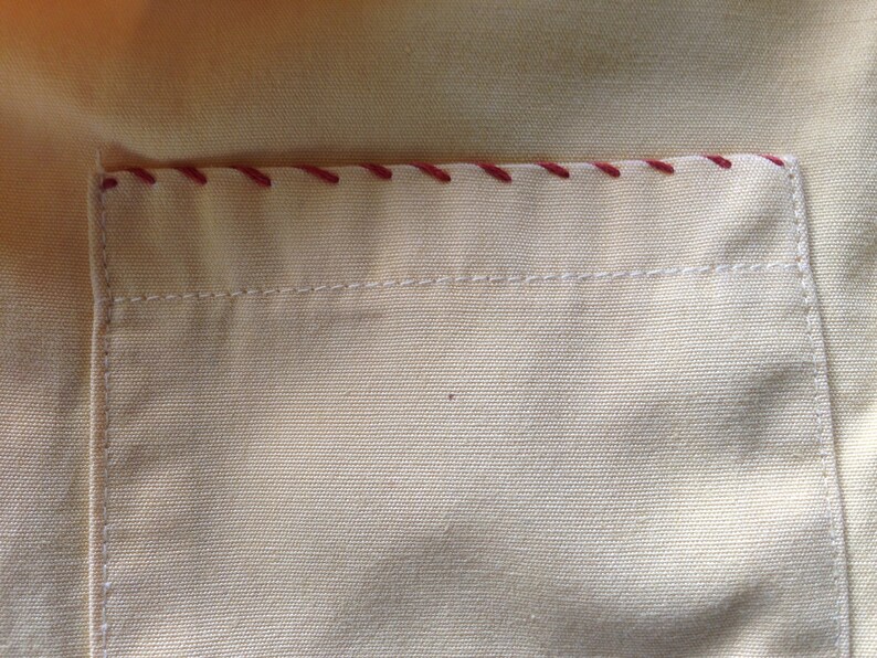 May include: A close-up of a beige fabric pocket with a red stitched border. The pocket is sewn onto a larger piece of beige fabric.
