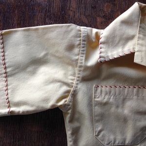 May include: A yellow short-sleeved shirt with red stitching around the collar, cuffs, and pocket. The shirt has a single pocket on the left chest.