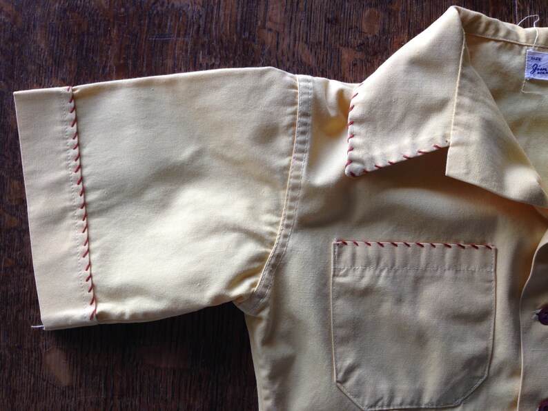 May include: A yellow short-sleeved shirt with red stitching around the collar, cuffs, and pocket. The shirt has a single pocket on the left chest.