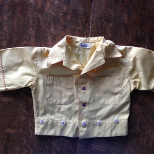 May include: A yellow short-sleeved button-up shirt with red stitching and white buttons. The shirt has a pocket on the left side and a label that reads "The Wee One".