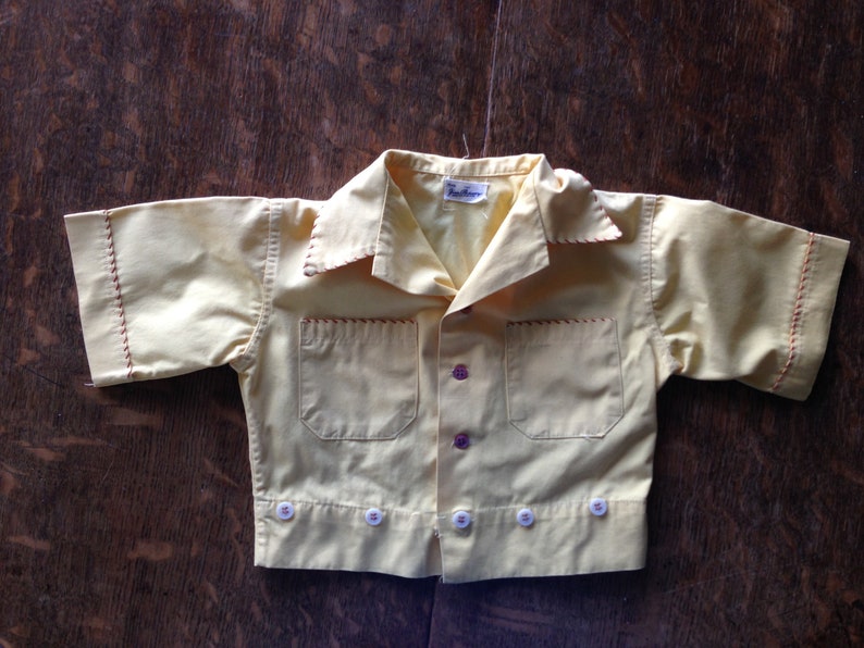 May include: A yellow short-sleeved button-up shirt with red stitching and white buttons. The shirt has a pocket on the left side and a label that reads "The Wee One".