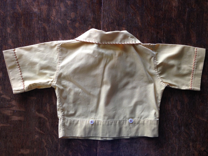 May include: A yellow short-sleeved shirt with a collar and two buttons on the bottom. The shirt has red stitching around the edges.