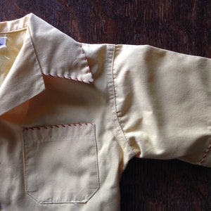 May include: A yellow short-sleeved shirt with red stitching around the collar, pocket, and sleeves. The shirt has a label that reads "Jim Palmer".