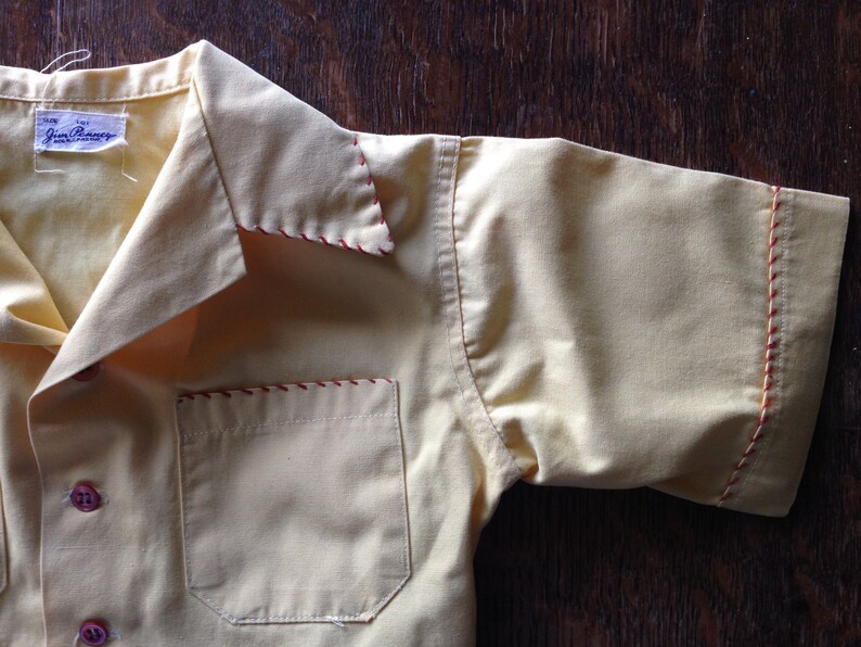 May include: A yellow short-sleeved shirt with red stitching around the collar, pocket, and sleeves. The shirt has a label that reads "Jim Palmer".