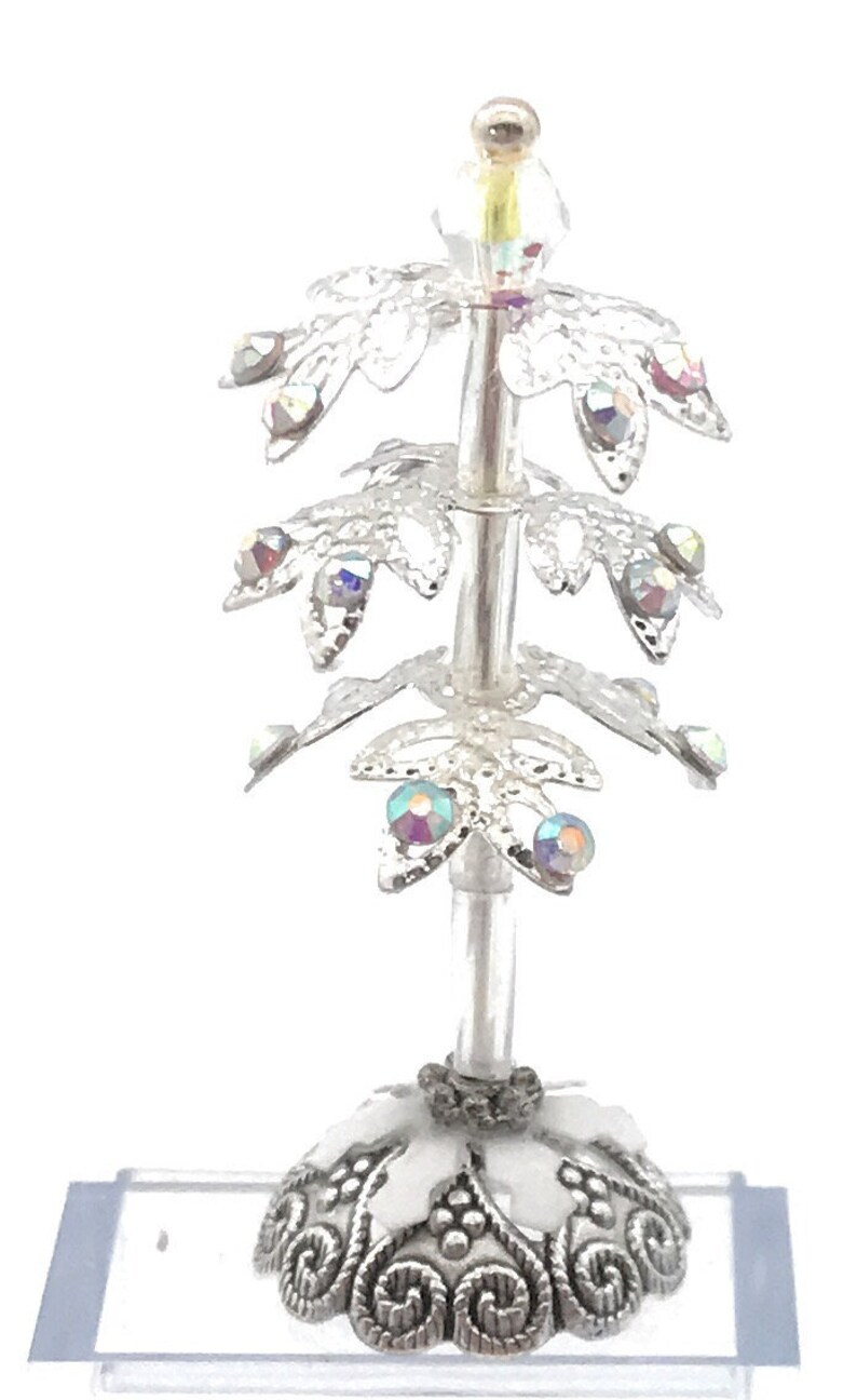 以下が含まれることがあります： 虹色のクリスタルが付いた銀と白のミニチュアクリスマスツリーのオーナメント。ツリーは渦巻き模様の装飾的なベースを持っています。