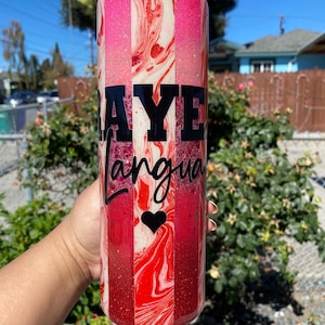 May include: A tall cylindrical tumbler featuring vertical stripes of glittery pink, red, and white marble patterns. The tumbler displays the black text "RAYE" in a bold sans-serif font above the cursive word "Langua" and a small black heart icon.