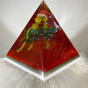 Pode incluir: Uma pirâmide de vidro vermelha com uma figura verde e amarela do Grinch segurando uma árvore de Natal dentro. O Grinch está usando um chapéu de Papai Noel vermelho.