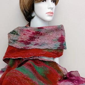 May include: A felted scarf featuring a textured, multi-colored design with vibrant shades of red, deep green, and muted purple. The fabric displays a layered, organic pattern with visible fiber textures throughout, draped around a mannequin neck.