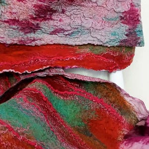 May include: A felted scarf featuring a textured surface with a blend of vibrant colors including deep red, magenta, forest green, and muted teal. The fabric displays layered, wavy patterns and organic, uneven edges throughout the material.