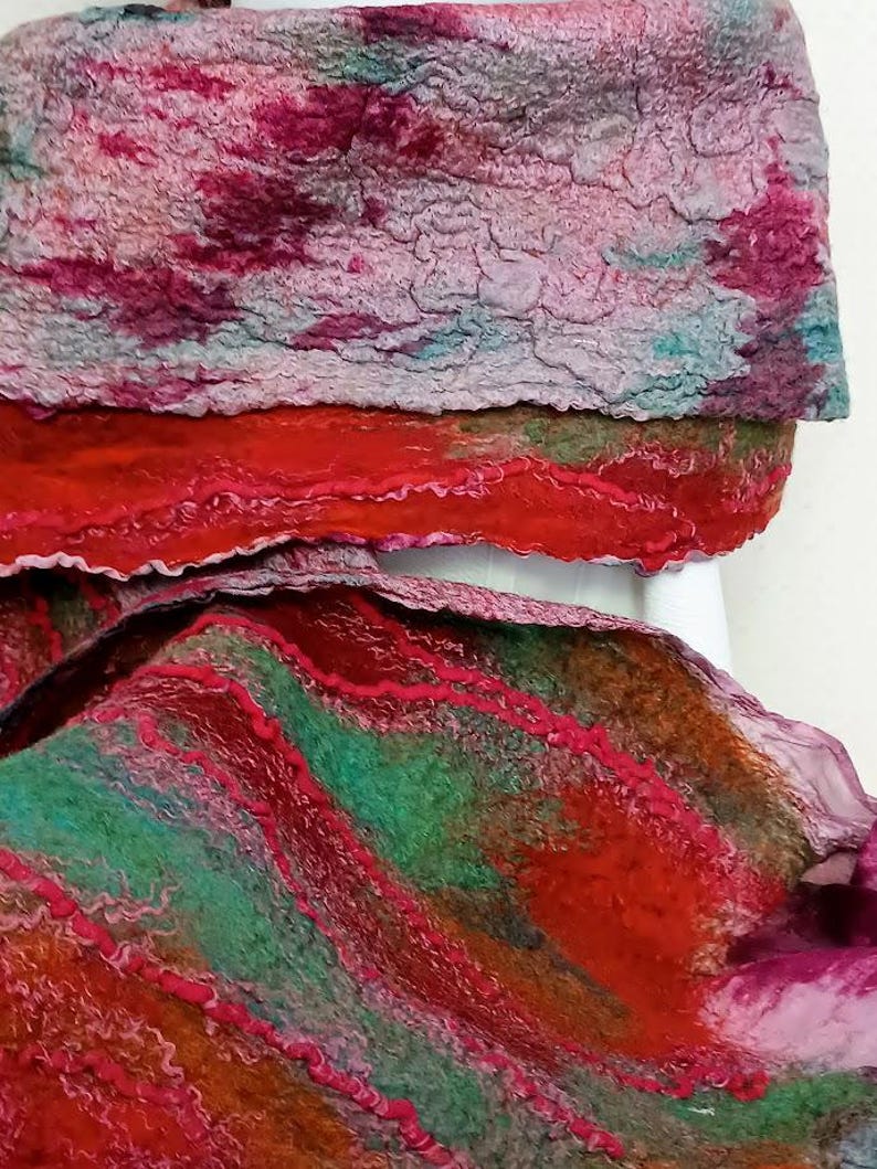 May include: A felted scarf featuring a textured surface with a blend of vibrant colors including deep red, magenta, forest green, and muted teal. The fabric displays layered, wavy patterns and organic, uneven edges throughout the material.