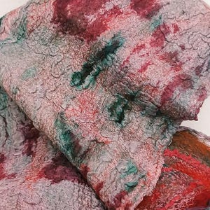 May include: A textured felted scarf featuring a mottled pattern of deep red, forest green, and muted gray tones. The fabric has a dense, uneven surface with visible fiber layering and a folded drape that reveals a vibrant red edge detail.
