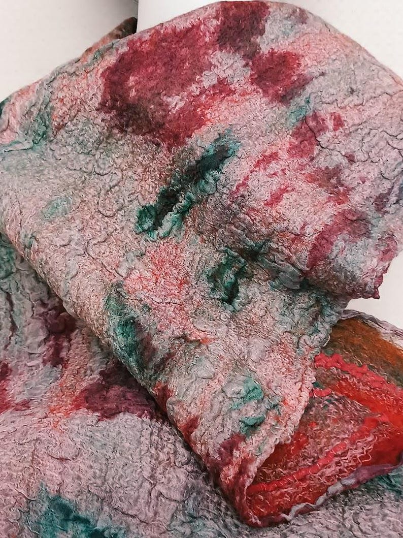 May include: A textured felted scarf featuring a mottled pattern of deep red, forest green, and muted gray tones. The fabric has a dense, uneven surface with visible fiber layering and a folded drape that reveals a vibrant red edge detail.