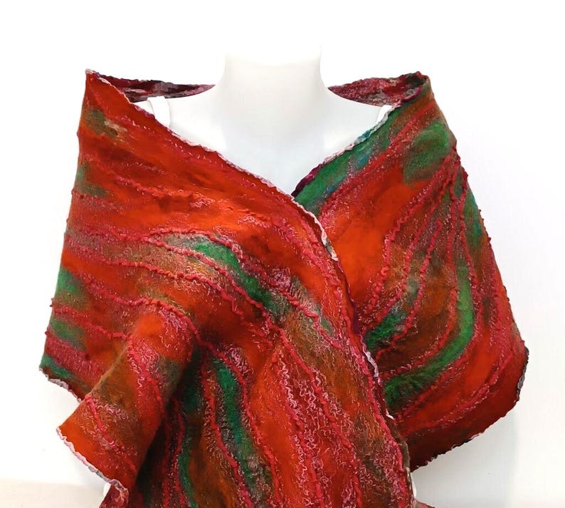 May include: A textured shawl featuring a vibrant pattern of diagonal stripes in shades of deep red, burnt orange, and forest green. The fabric has a thick, felted appearance with raised, wavy ridges running across the surface of the garment.