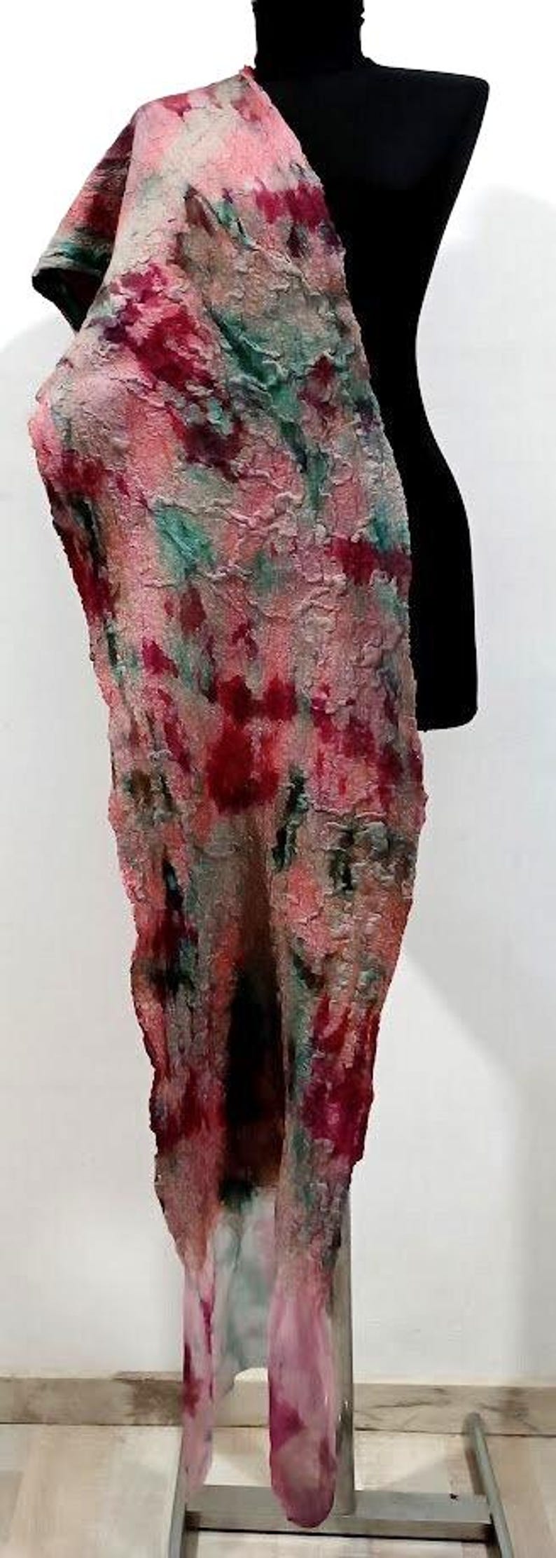 May include: A long, textured scarf featuring a mottled pattern of pink, deep red, and teal green hues. The fabric has a felted, uneven surface with visible fibers and organic color blending, draped vertically over a black display form.