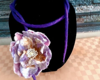 Puede incluir: Una flor de fieltro morado y blanco con un centro de pedrería, unida a un collar de cordón morado.
