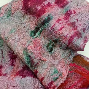 May include: A felted scarf featuring a textured, mottled surface with a blend of deep magenta, soft pink, muted gray, and forest green hues. The fabric displays a dense, fibrous appearance with irregular patterns and a folded, layered structure.