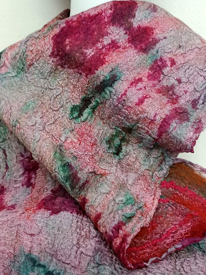 May include: A felted scarf featuring a textured, mottled surface with a blend of deep magenta, soft pink, muted gray, and forest green hues. The fabric displays a dense, fibrous appearance with irregular patterns and a folded, layered structure.