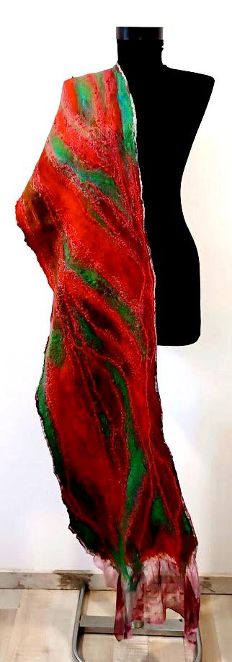 May include: A long, rectangular felted scarf featuring a vibrant, swirling pattern of deep red, bright orange, and forest green fibers. The textile has a textured, organic surface with uneven edges and a sheer, ruffled hem at the bottom end.