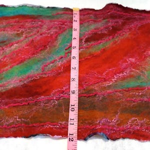 May include: A felted textile piece featuring blended shades of red, orange, and teal with wavy fiber textures. A pink measuring tape is placed vertically across the center, showing a width of approximately 14 inches for the fabric section.