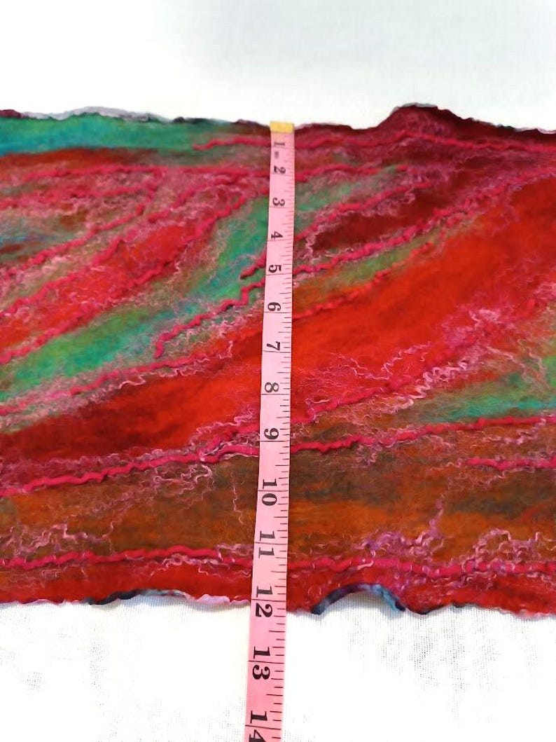 May include: A felted textile piece featuring blended shades of red, orange, and teal with wavy fiber textures. A pink measuring tape is placed vertically across the center, showing a width of approximately 14 inches for the fabric section.