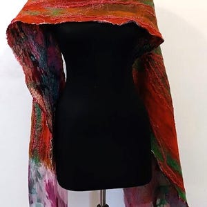 May include: A long, textured scarf featuring a vibrant blend of orange, red, green, and purple hues. The fabric displays a layered, felted appearance with raw, uneven edges and a flowing, lightweight drape that transitions into a sheer, dyed finish.
