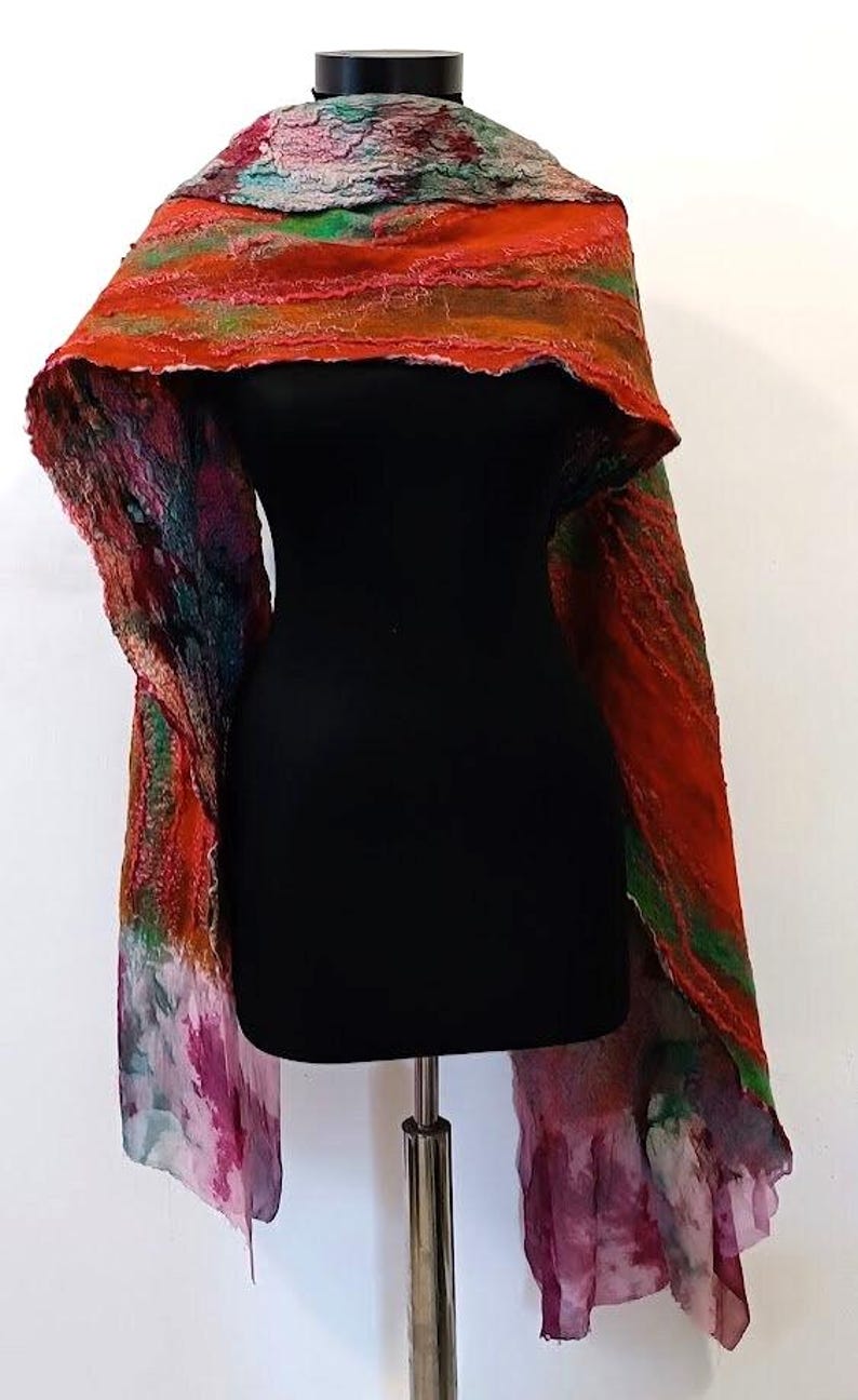 May include: A long, textured scarf featuring a vibrant blend of orange, red, green, and purple hues. The fabric displays a layered, felted appearance with raw, uneven edges and a flowing, lightweight drape that transitions into a sheer, dyed finish.