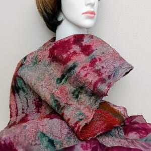 May include: A textured felted scarf featuring a mottled pattern of deep red, muted green, and grey tones. The scarf has a thick, uneven surface texture and is layered over a sheer, lightweight fabric with a matching dark pink and green abstract print.