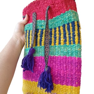 May include: A colorful woven backpack with a drawstring closure and purple tassels. The backpack is made of a variety of colors, including red, pink, yellow, green, and blue. The text "Handmade Marcella" is visible on the bottom of the backpack.
