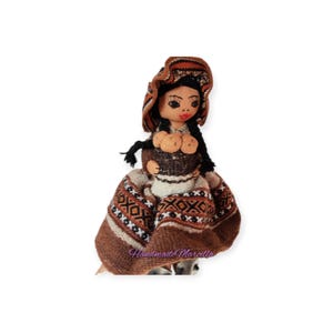 May include: Handmade doll with a traditional design. The doll wears a brown and white patterned dress and a matching hat. The doll has black braids and a painted face with red lips. The doll is handmade and has the text "Handmade Marcella" on the dress.