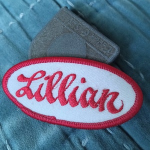 以下が含まれることがあります： 青い布地に、赤い筆記体で「Lillian」と刺繍された白い楕円形のワッペン