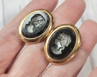 Könnte beinhalten: Ein Paar ovale Ohrringe mit goldfarbenem Rand. Jeder Ohrring hat ein schwarzes Cameo mit einem weißen Profil einer Frau. Die Ohrringe sind vor einem hellen Hintergrund platziert.