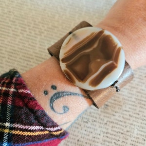 May include: A brown leather bracelet with a large, round, white and brown agate stone. The bracelet is worn on a person's wrist.